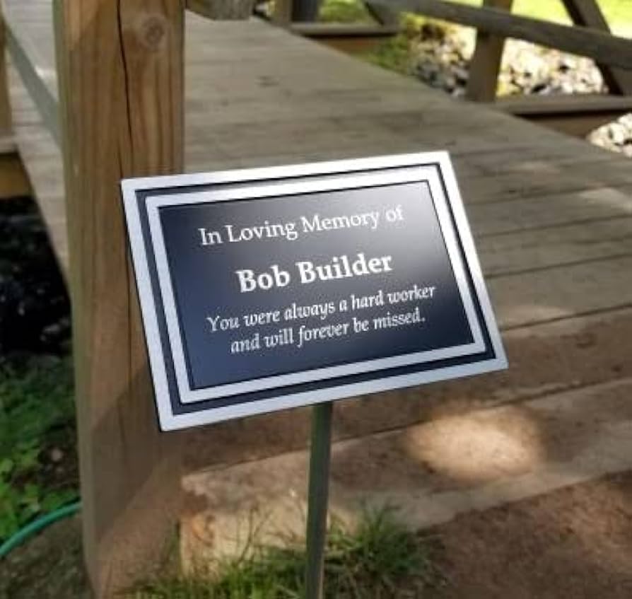 Cast aluminum memorial plaque with black background, silver border, and white text on a stake.