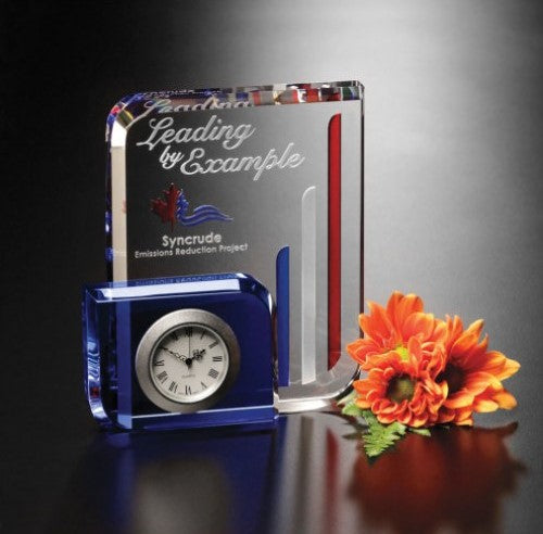 Glass desk clock with blue block accent, silver face, black Roman numerals, beside an etched crystal award.