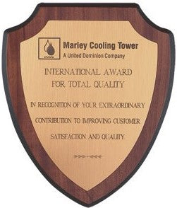 Shield-shaped wood plaque with a bright gold reflective plate, engraved with text and a logo.